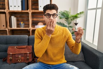 Young hispanic man holding sand clock covering mouth with hand, shocked and afraid for mistake....