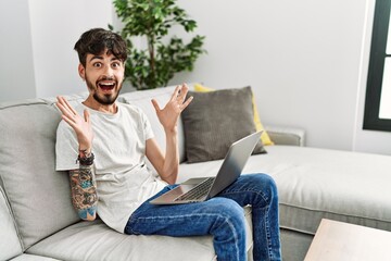 Hispanic man with beard sitting on the sofa celebrating crazy and amazed for success with arms raised and open eyes screaming excited. winner concept