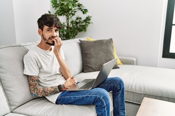 Fototapeta premium Hispanic man with beard sitting on the sofa looking stressed and nervous with hands on mouth biting nails. anxiety problem.