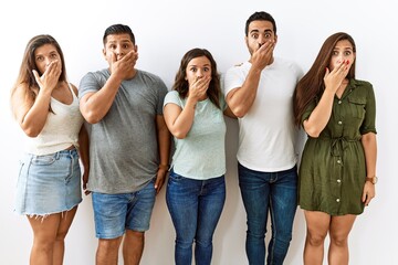 Group of young hispanic friends standing together over isolated background shocked covering mouth with hands for mistake. secret concept.