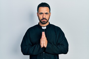 Handsome hispanic priest man with beard standing over isolated background skeptic and nervous,...