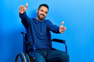 Handsome hispanic man with beard sitting on wheelchair looking at the camera smiling with open arms for hug. cheerful expression embracing happiness. - obrazy, fototapety, plakaty