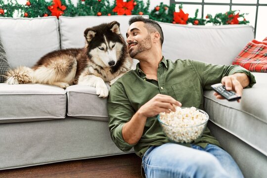 Young hispanic man watching movie sitting on sofa with dog by christmas decor at home