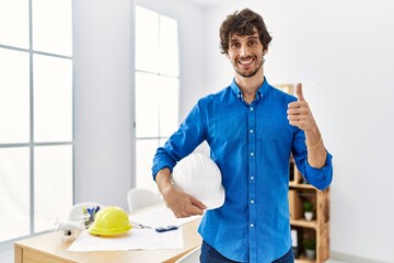 Young hispanic man with beard holding safety helmet and blueprints at the office smiling happy and positive, thumb up doing excellent and approval sign