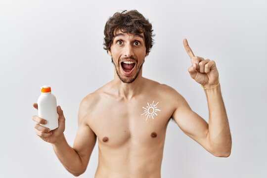 Young hispanic man standing shirtless holding sunscreen lotion smiling amazed and surprised and pointing up with fingers and raised arms.