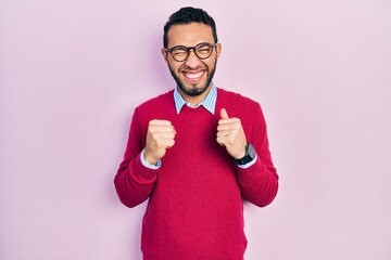 Hispanic man with beard wearing business shirt and glasses excited for success with arms raised and...