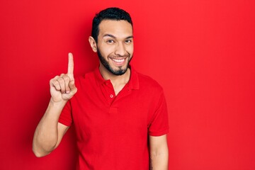 Hispanic man with beard wearing casual red t shirt showing and pointing up with finger number one while smiling confident and happy.