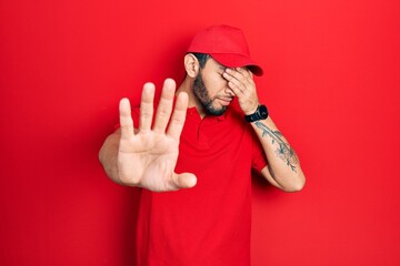 Hispanic man with beard wearing delivery uniform and cap covering eyes with hands and doing stop gesture with sad and fear expression. embarrassed and negative concept.