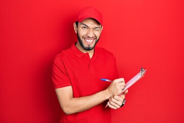 Hispanic man with beard wearing courier uniform holding clipboard winking looking at the camera with sexy expression, cheerful and happy face.