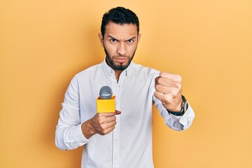 Hispanic man with beard holding reporter microphone annoyed and frustrated shouting with anger, yelling crazy with anger and hand raised