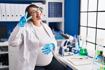 Pregnant woman working at scientist laboratory doing peace symbol with fingers over face, smiling cheerful showing victory