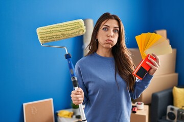Young brunette woman holding roller painter painting new house puffing cheeks with funny face....