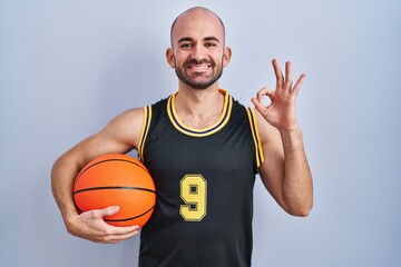 Young bald man with beard wearing basketball uniform holding ball smiling positive doing ok sign with hand and fingers. successful expression.