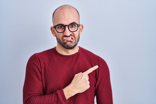 Young bald man with beard standing over white background wearing glasses pointing aside worried and nervous with forefinger, concerned and surprised expression