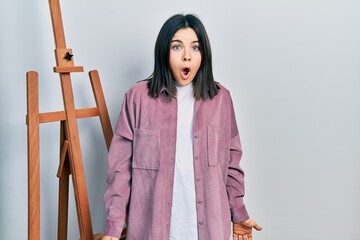 Young brunette woman standing by empty easel stand scared and amazed with open mouth for surprise,...