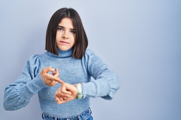 Young hispanic woman standing over blue background in hurry pointing to watch time, impatience,...