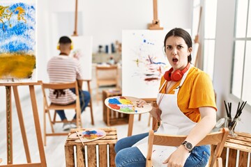 Young artist woman painting on canvas at art studio in shock face, looking skeptical and sarcastic, surprised with open mouth