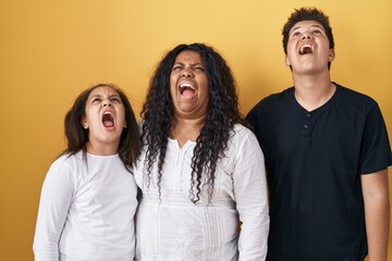 Family of mother, daughter and son standing over yellow background angry and mad screaming frustrated and furious, shouting with anger. rage and aggressive concept.