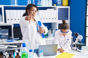 Mother and young daughter working at scientist laboratory pointing with finger to the camera and to...