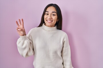 Young south asian woman standing over pink background showing and pointing up with fingers number three while smiling confident and happy.