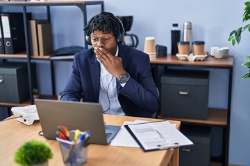 Young african man with dreadlocks working at the office wearing headset covering mouth with hand,...