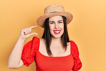 Young hispanic woman wearing summer hat smiling and confident gesturing with hand doing small size sign with fingers looking and the camera. measure concept.