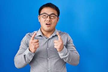 Young chinese man standing over blue background amazed and surprised looking up and pointing with fingers and raised arms.