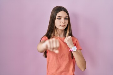 Teenager girl standing over pink background punching fist to fight, aggressive and angry attack,...