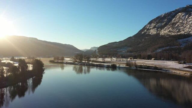 Beautiful vibrant winter morning sunrise above Hallingdal river in Nesbyen Norway - Aerial flying forward above river surface with sun coming in to frame from left