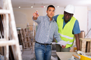 Caucasian man architect and african-american man engineer having conversation about work plan in apartment. Pointing with finger, showing with hand gesture.