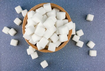 Scattered sugar cubes in a wooden bowl on a blue background. Sugar in a wooden carved bowl on a...