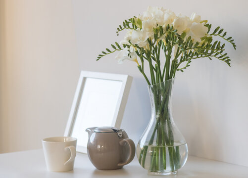 Closeup of white freesia flowers with teapot, cup and small picture frame in background (selective focus)