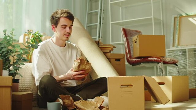 Moving to new house. Real estate, buying flat apartment. Young man wrapping cup in cardboard boxes, guy owner during relocation. Property purchasing. Housing service concept.