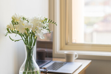 Closeup of white freesia flowers in glass vase with computer on desk in background next to window...