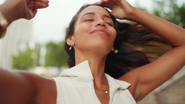 Close-up of smiling cute tanned woman with long brown hair wearing white top raises her hands and touches her hair. The girl enjoys the breeze. Camera moving forwards     