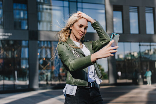 Young hipster Italian girl making selfie touching blonde hair against modern building. Beautiful young woman walking, making video call, swedish student girl on vacations, traveling having fun outdoor