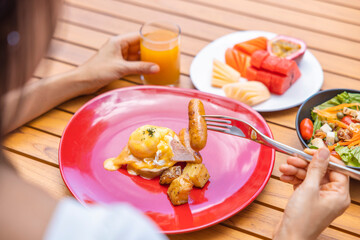 Woman's hand holding a fork and eating Breakfast. Egg Benedict, fruits such as watermelon, papaya, melon, passion fruit, orange juice and coffee. placed on a gray placemat