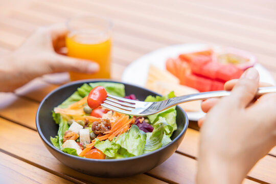 Woman's Hand Holding A Fork And Eating Breakfast. Vegetable Salad, Fruits Such As Watermelon, Papaya, Melon, Passion Fruit, Orange Juice And Coffee. Placed On A Gray Placemat