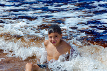 Happy boy splashing in the waves of the Baltic Sea on a bright summer day in Jurmala, Latvia. Summertime and swimming activities for happy children.