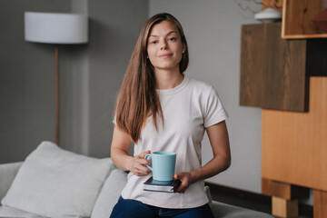 Confident European young adult woman in casual holding a cup of coffee putted on her diary. Pretty hispanic female standing at living room looking at camera. Remote employee on break. Distant work.