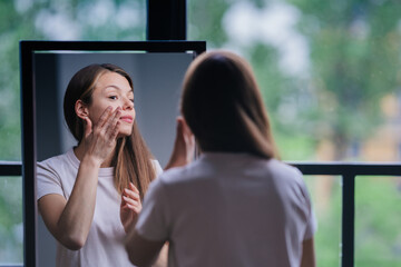 Young brunette woman in white t-shirt standing at mirror touching face at window with backdrop of blurry green trees. Beautiful girl having morning body care. Confident housewife at home. Healthy life