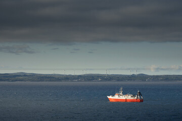 Fototapeta premium Small fishing ship with red hull in the ocean. Galway bay, Ireland. Food industry and supply concept. Cloudy sky and dark color water surface.