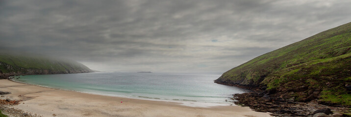 Panorama image of Keem bay and beach. Achill island, county Mayo, Ireland. Low clouds over ocean and mountains. Popular tourist area with stunning nature scenery and clean blue water and sandy beach