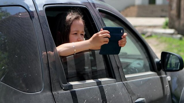 Portrait of cute child girl shoots photo or video on her smart phone while traveling, from car window 4k