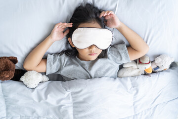 Top view child little asian girl blindfold sleeps in the bed with a toy teddy bear