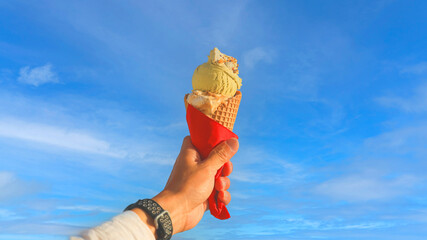 Man hand holding delicious ice cream with macadamia nuts and vanilla waffle cone with blue sky...