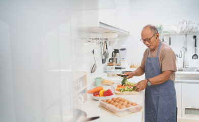 Happy retired Asian Mature adult man standing chopping fresh broccoli on wooden board while preparing Healthy food, Senior Male cooking in Modern kitchen.