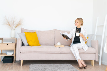 Middle age woman working at home in business suit sitting on couch using reading notebook looks at plans for the day and drinking morning coffee. Caucasian business woman in the living room