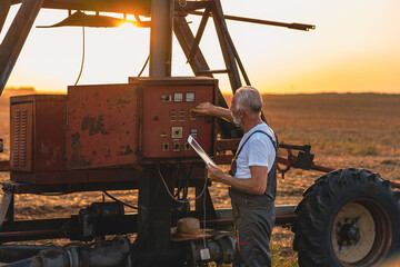 Older farmer holding tablet in his hands and adjusts irrigation system on field at sunset.