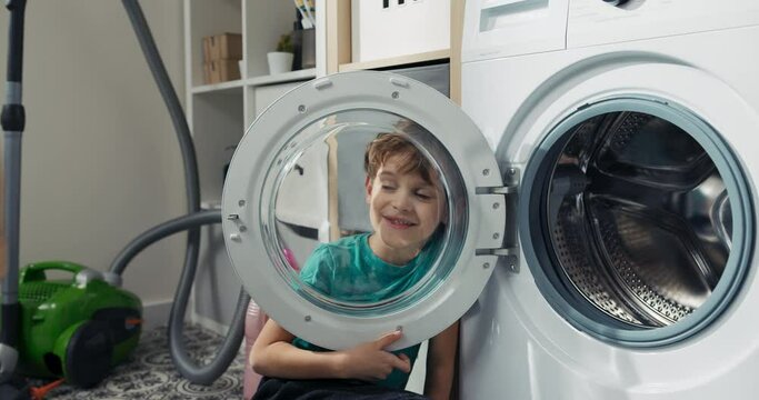 Smiling boy with dark eyes looks through round glass door of washing machine, fools around while doing household chores.